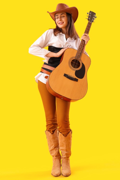 A woman wears brown cowboy boots with white top and a wide brimmed hat and is holding a guitar