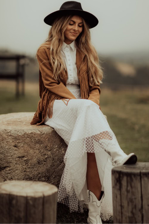 A woman wears cowboy boots with a long white dress, fringe coat and a cowboy hat
