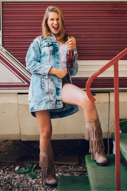 A woman wears fringe cowboy boots with with denim shorts, denim coat and a white top