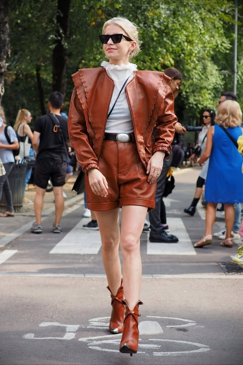 a woman wears brown outfit with jackets, leather shorts and ankle cowboy boots