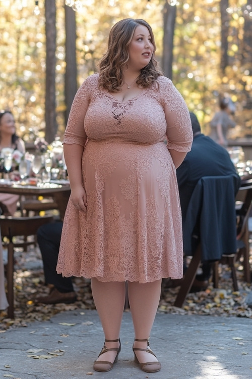 A plus-size woman wears a blush pink lace wedding guest dress with brown flats