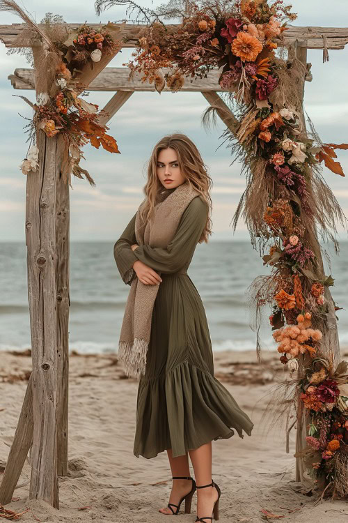 A woman in a layered olive green midi dress with a cozy scarf and block heels, posing near a wooden archway decorated with autumnal flowers for a beach wedding (2)