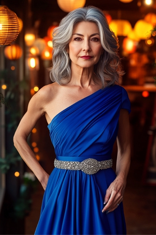 A woman over 50 wears a blue on shoulder wedding guest dress with a silver belt