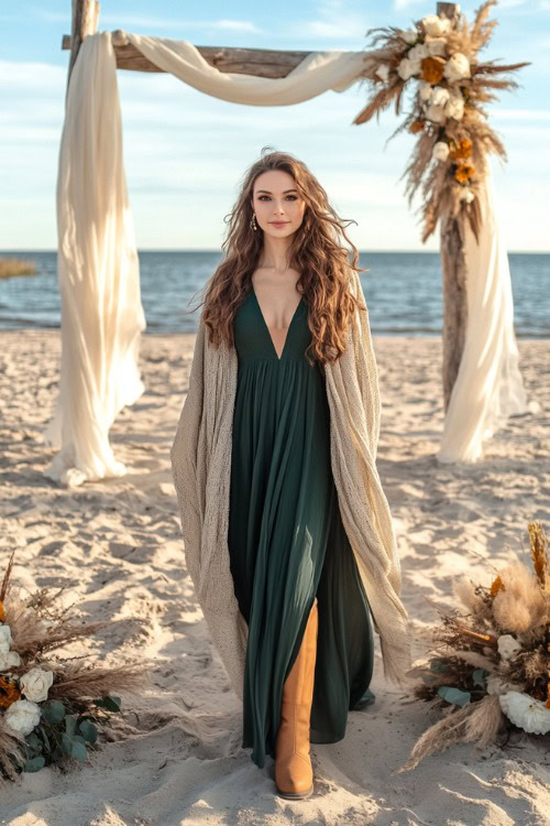 Layered fall look with a flowy dark green dress and a wraparound shawl, paired with tan boots on a sandy beach with a wedding arch in the background