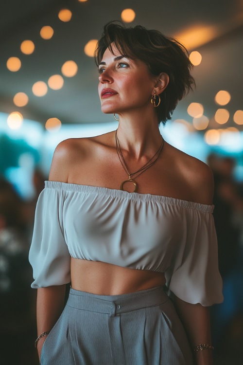 A mature woman in a stylish off-shoulder blouse tucked into high-waisted wide-leg trousers, paired with pointed-toe pumps and delicate jewelry