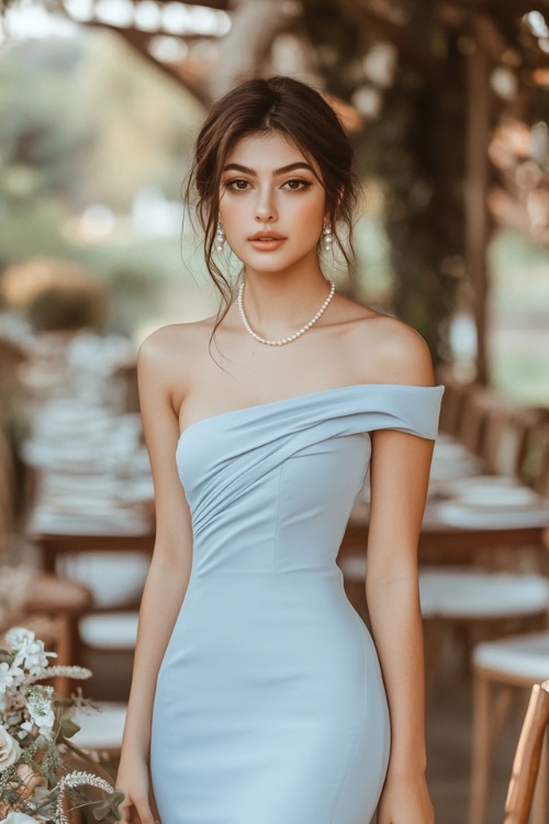 A woman in a powder blue one-shoulder dress with an asymmetrical hem, paired with delicate pearl accessories, posing by an outdoor wedding setup with simple wooden chairs