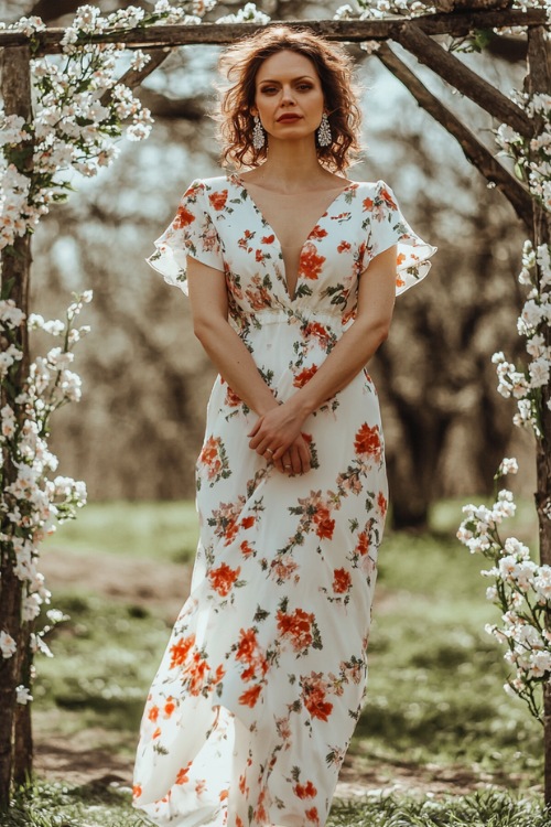 A stylish woman over 40 in a floral-printed chiffon maxi dress with short flutter sleeves and a flowing silhouette, accessorized with dainty earrings and sandals (3)